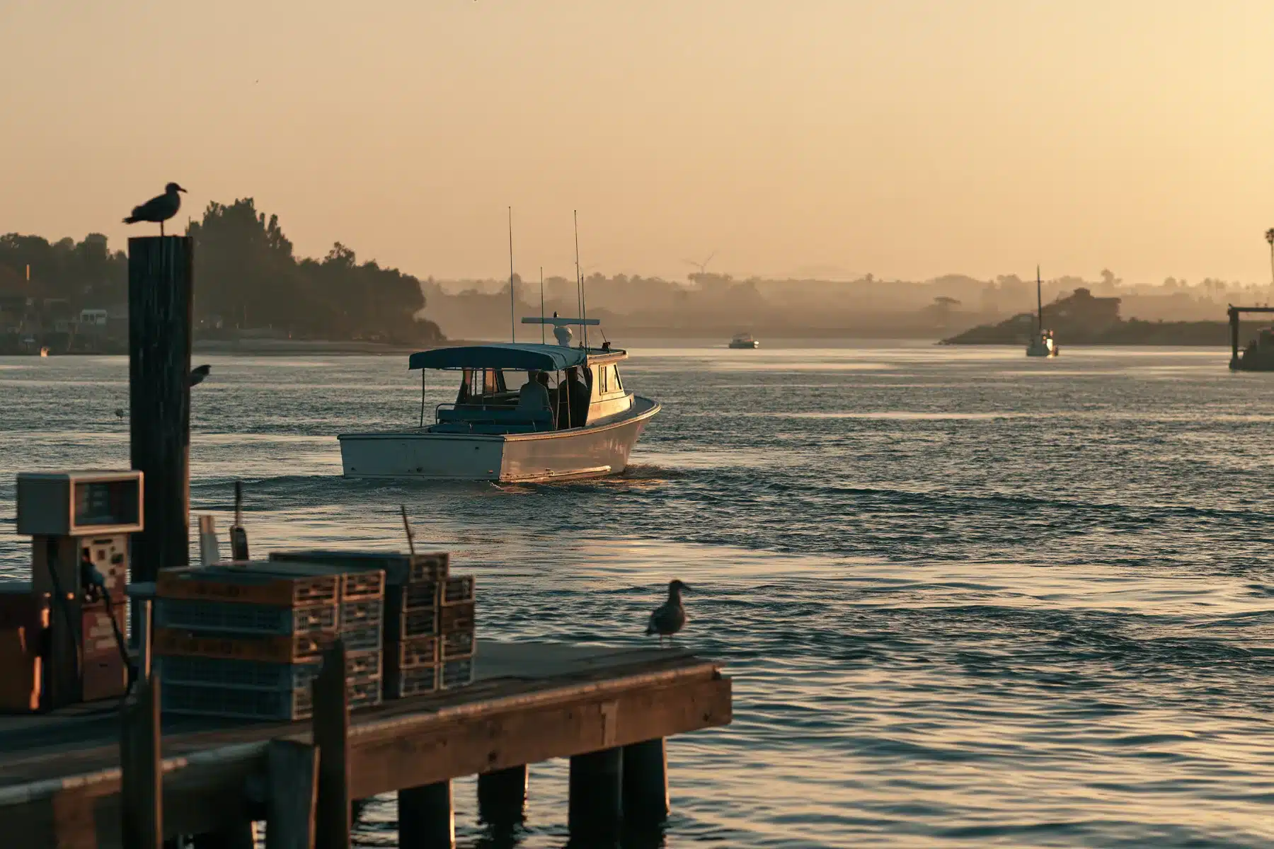 A working boat with a canopy heading out past a fuel dock and stacked crates in a harbor at first light