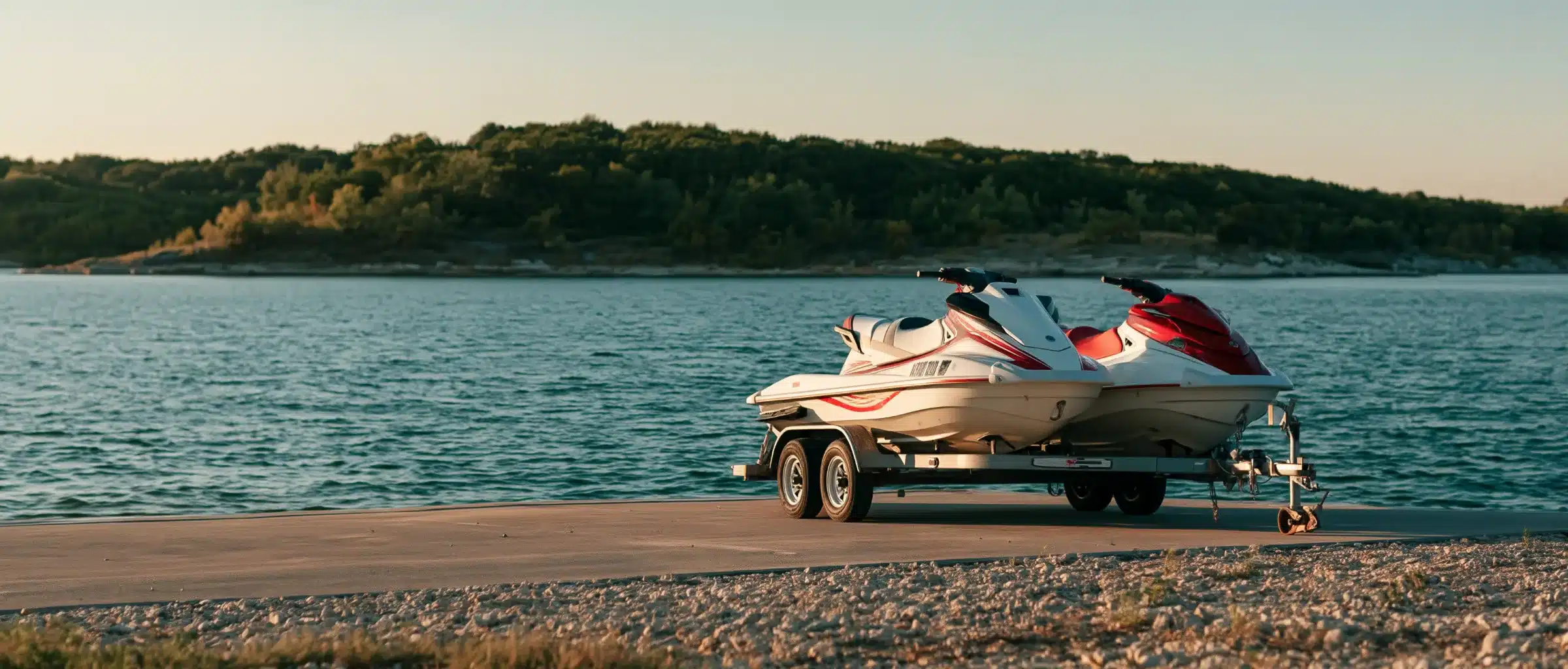 Two personal watercraft on a double trailer at a lake boat ramp in warm evening light