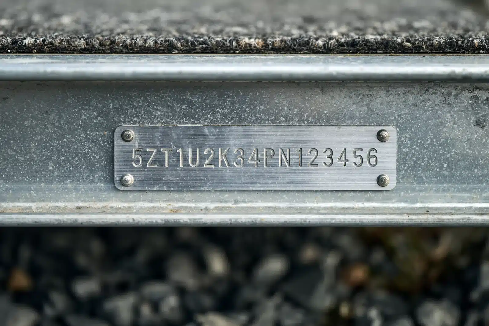 A brushed aluminum identification plate with a seventeen-character trailer number stamped on it, riveted to a galvanized steel boat trailer frame rail above gravel