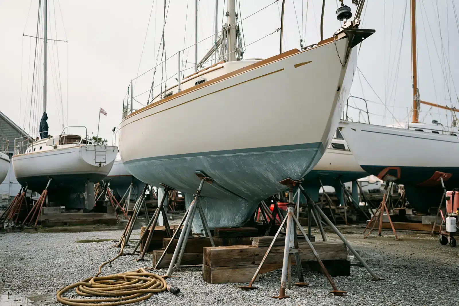 A cream sailboat with teak trim standing on steel jack stands and timber blocks in a boatyard, with its worn blue bottom paint showing and other sailboats on stands behind