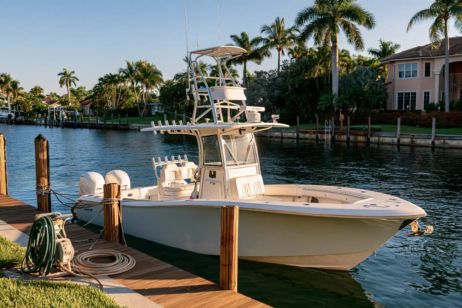 A white center console fishing boat with twin outboards, a T-top and rod holders tied alongside a wooden dock on a palm-lined canal at sunrise