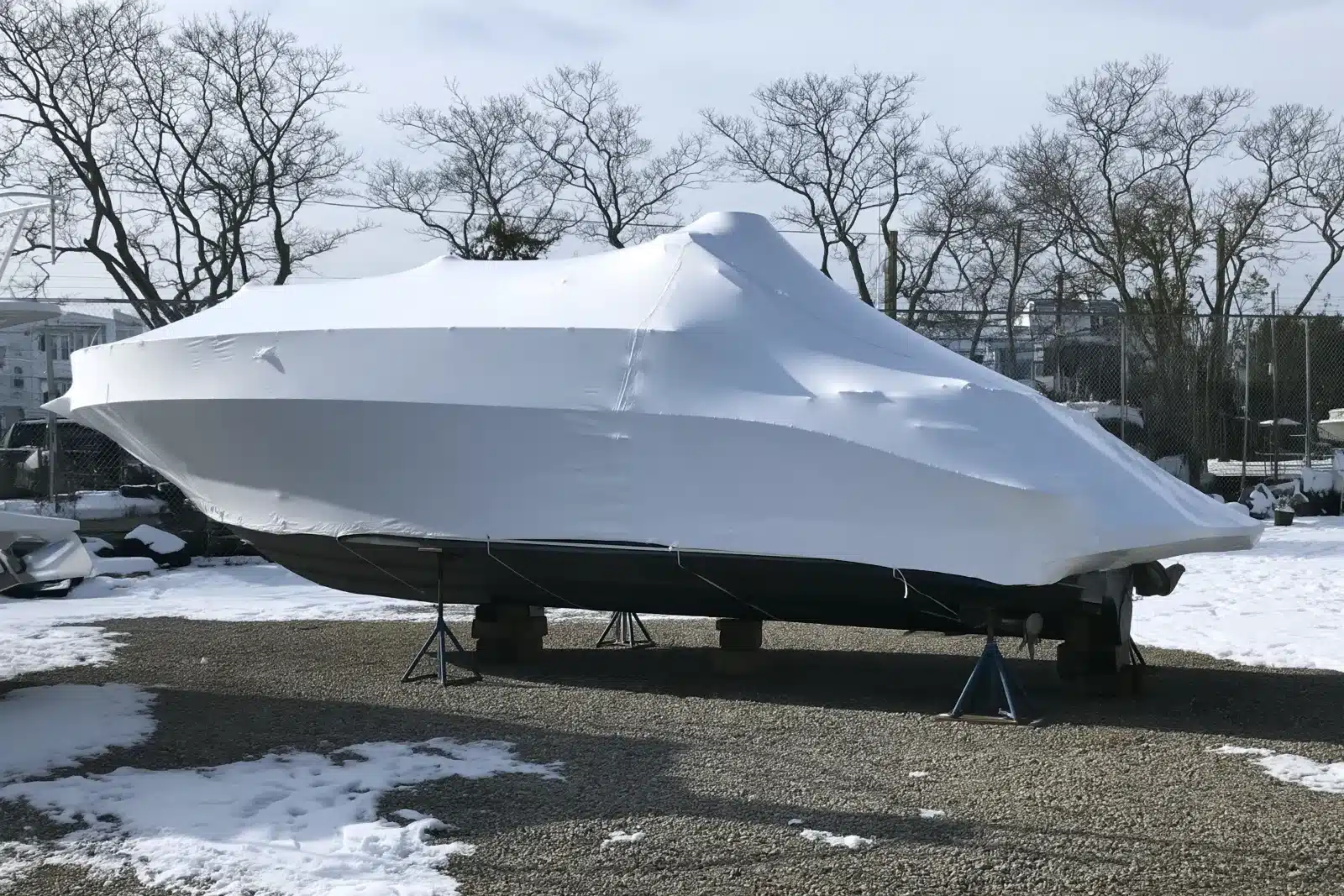 A powerboat shrink-wrapped in tight white plastic on jack stands and blocks in a snowy boatyard, with its dark bottom showing below the wrap and bare trees and a chain-link fence behind