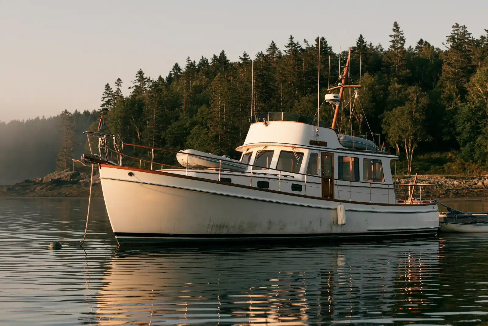 A classic white trawler with a teak-trimmed pilothouse and a dinghy on deck at anchor on calm water at sunrise, with a pine-covered shoreline behind