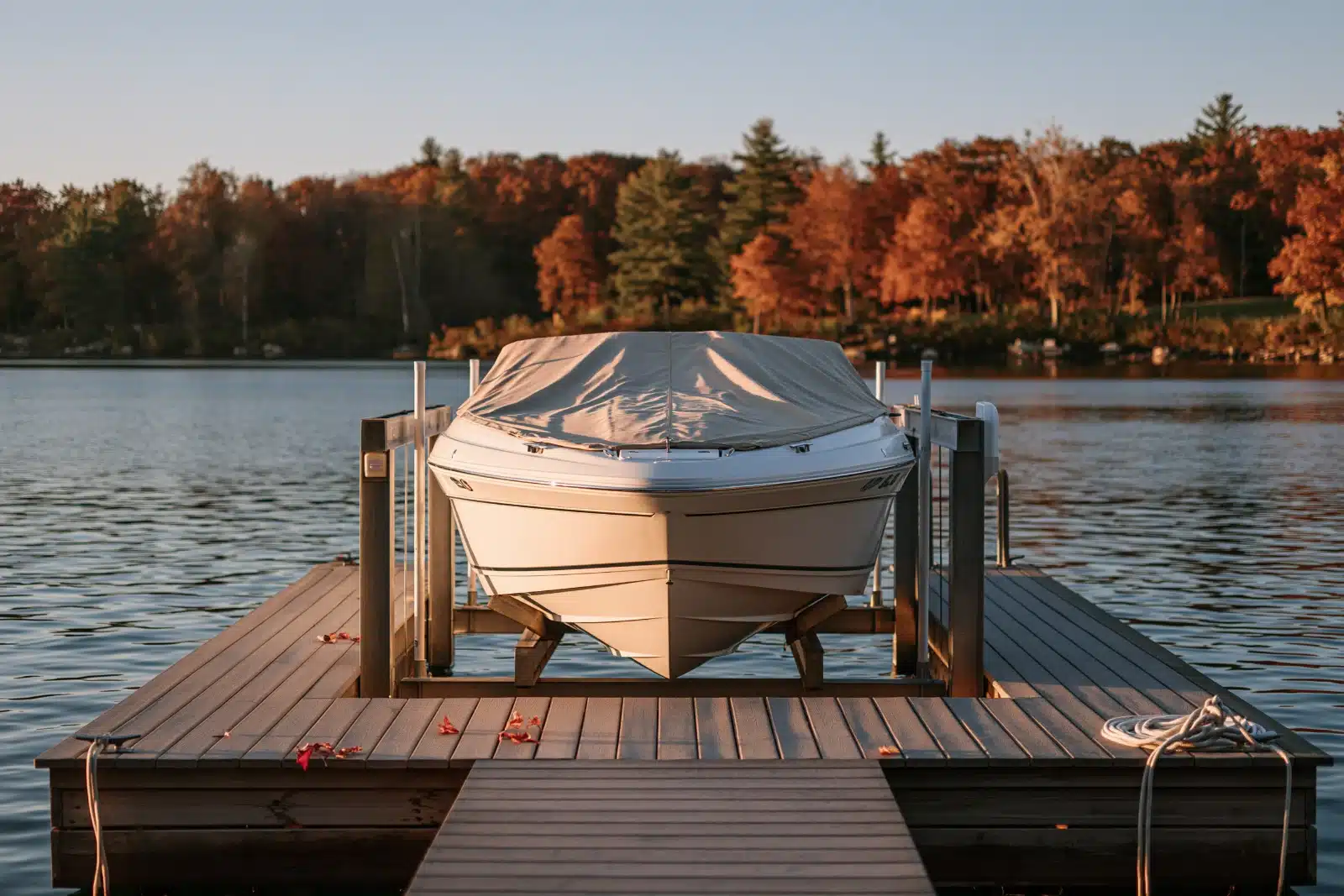 A covered powerboat raised out of the water on a boat lift at the end of a wooden lake dock in fall, with red and orange leaves on the planks, a coiled dock line and autumn trees across the water