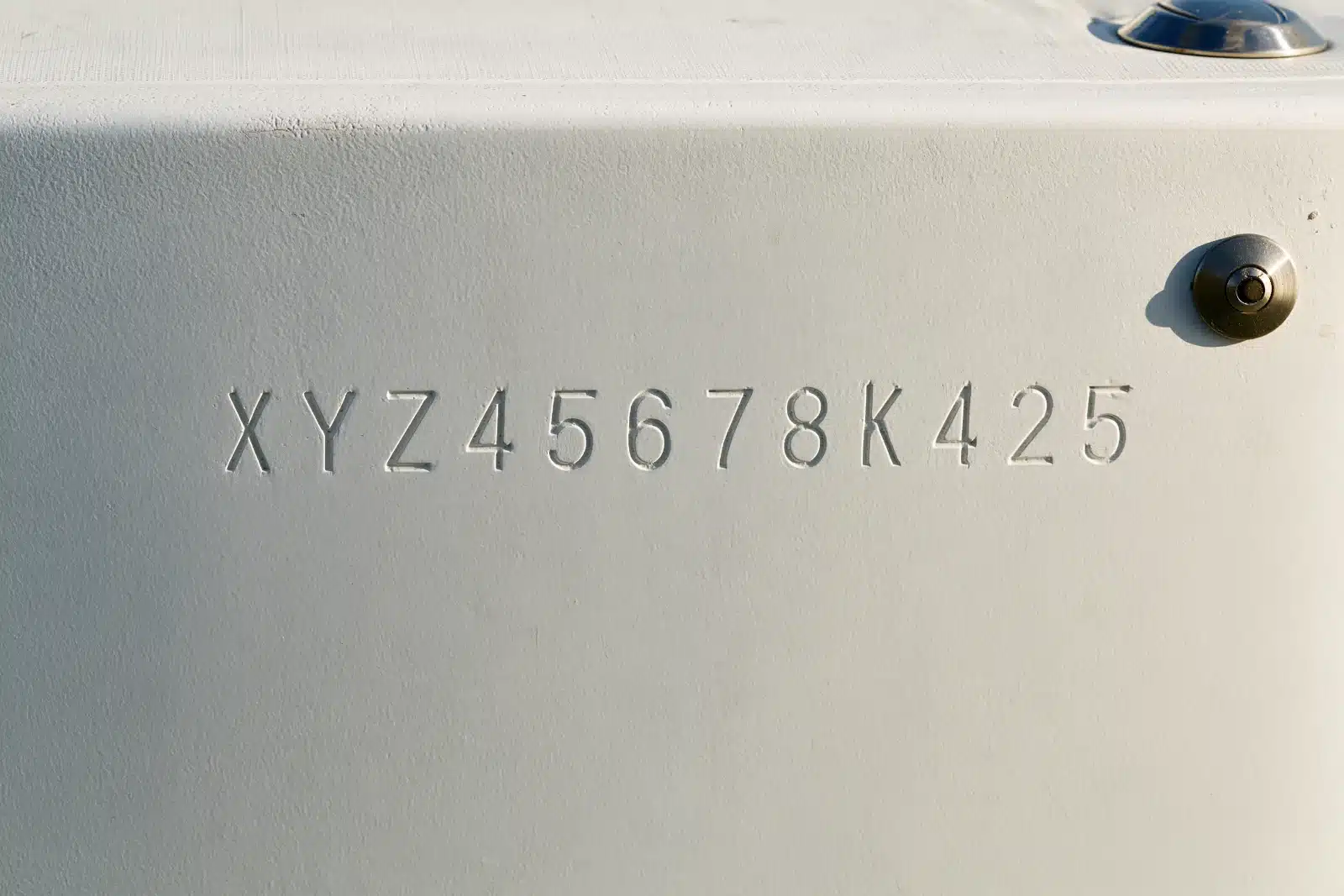 A close view of a white fiberglass boat transom with a twelve-character hull identification number engraved in a straight line into the gelcoat, and a stainless bolt head to the right