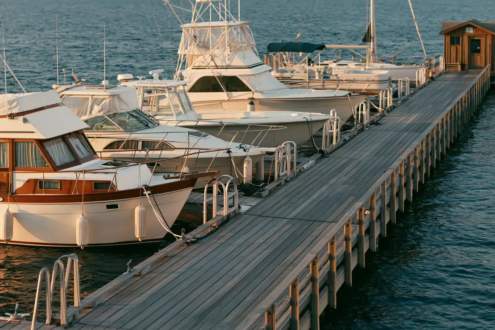 A row of used boats of different types tied alongside a long wooden dock at sunset, with fenders, boarding steps at the dock, and a small wooden office at the far end