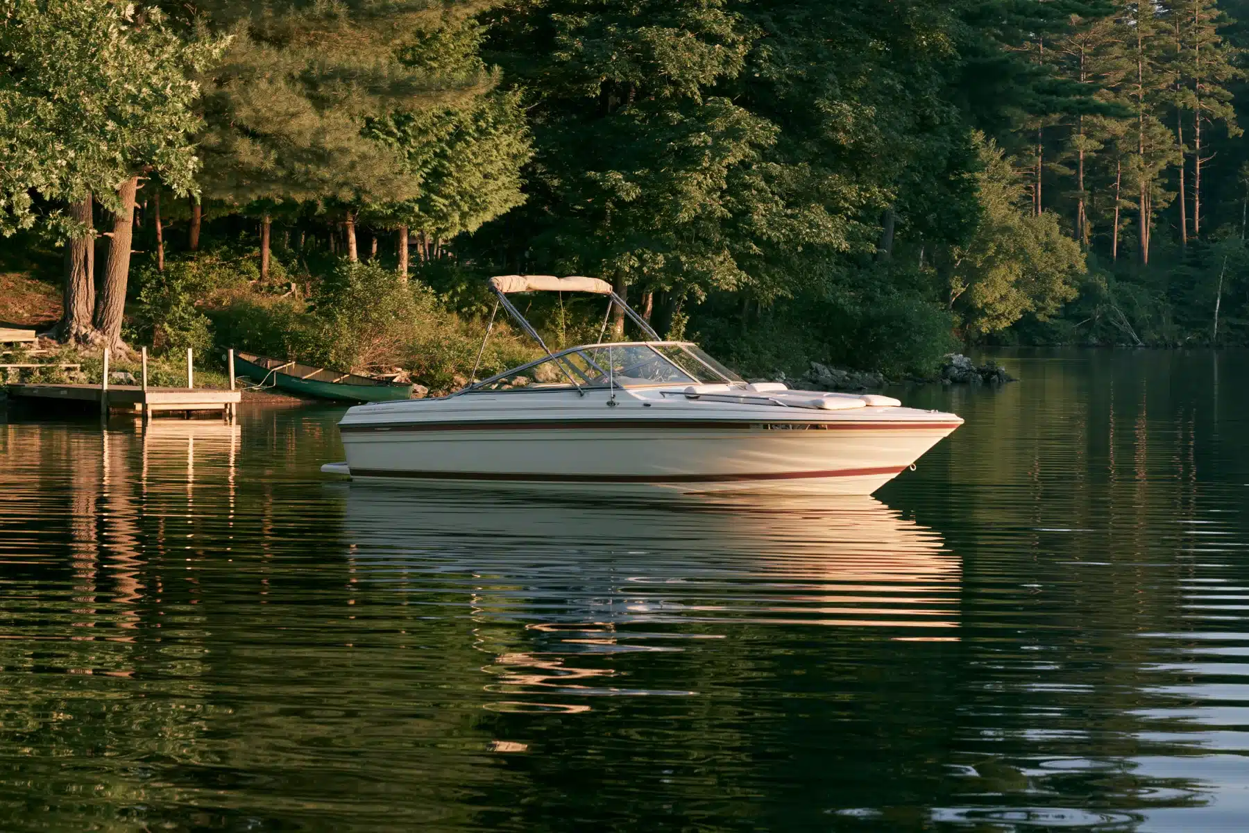 A cream bowrider with its bimini top up, idling on a calm wooded lake near a small dock