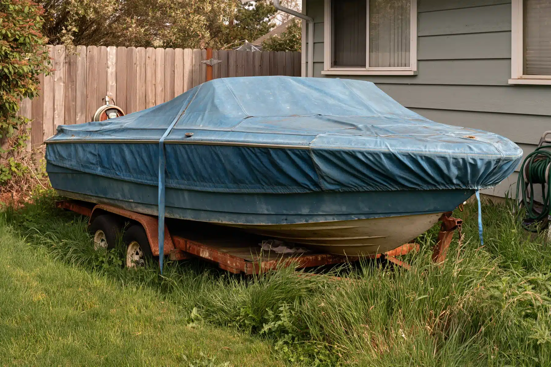 An older fiberglass runabout under a faded blue cover on a rusted trailer in the side yard of a ranch home
