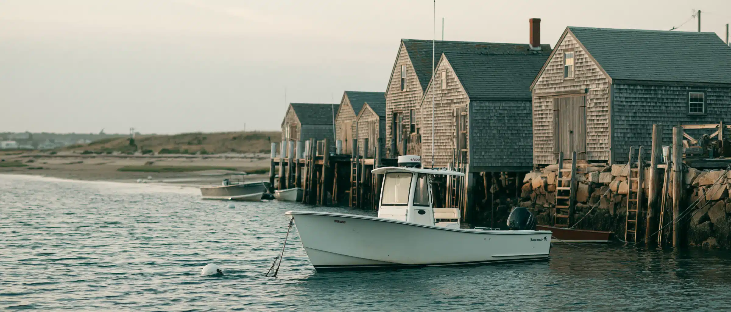 A white center console boat on a mooring in a Cape Cod harbor, with weathered gray-shingled fish shacks on pilings, a stone bulkhead and a dune-grass spit at dusk