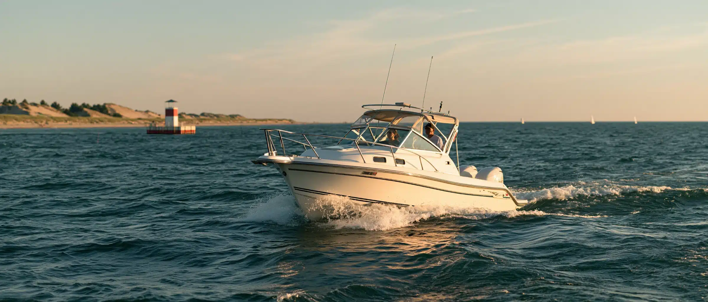 A white cabin cruiser running across blue Lake Michigan in evening light, with a red-and-white pier lighthouse, sand dunes and distant sailboats behind it