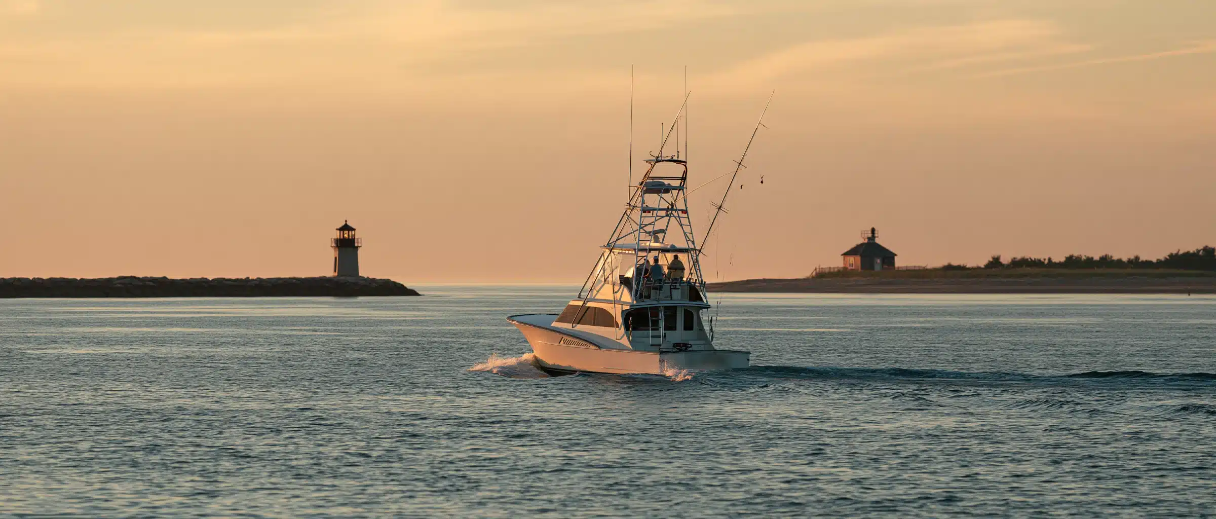 A tuna-tower sportfishing boat leaving an inlet at dusk on Long Island, past a jetty lighthouse and a dune cottage