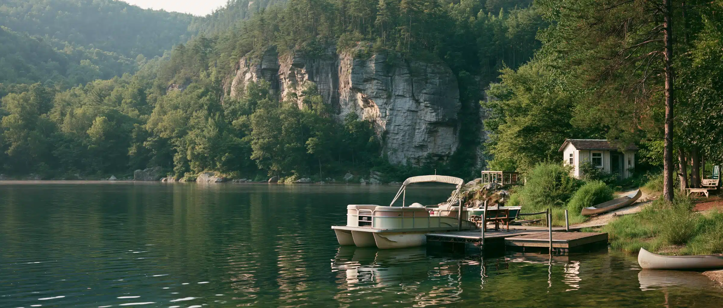A pontoon boat with a bimini top at a small wooden dock on a still green lake below a pale rock bluff, with forested hills, a white cabin and canoes on the shore