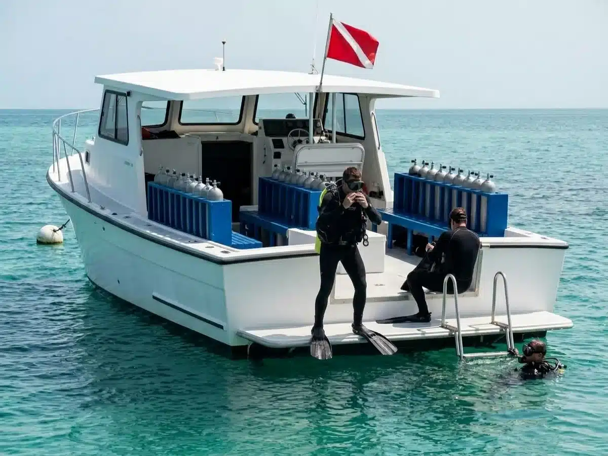 A white commercial dive charter boat on a mooring in turquoise water, tank racks on deck, one diver at the edge of the swim step about to stride in and another waiting at the ladder