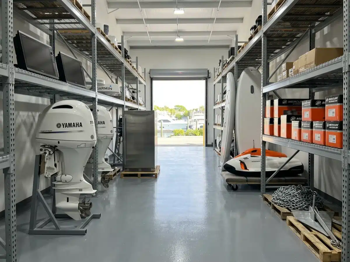 An aisle in a marine consignment warehouse stocked with white Yamaha outboards, Garmin displays and radar, a stainless galley fridge, a Seabob, paddleboards, boxed Mercury parts and anchor chain, the roll-up door open to a marina