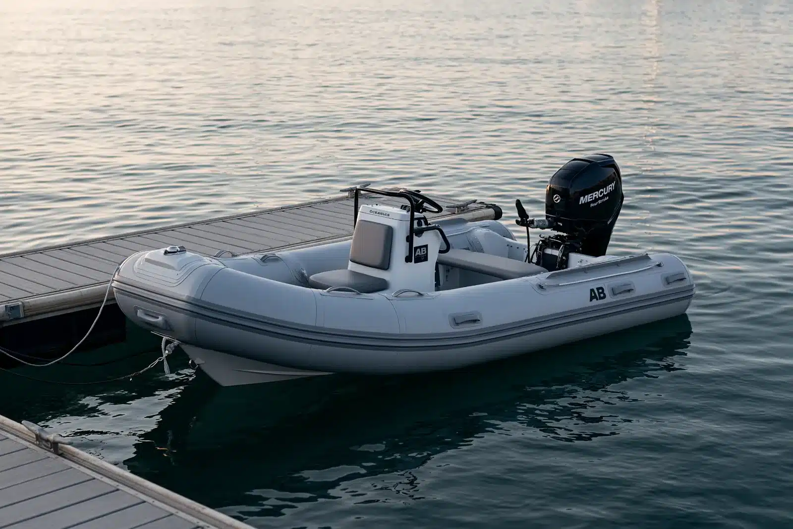 A gray AB rigid inflatable tender with a center console and a black Mercury outboard, tied by its bow line to a floating dock on calm water at dusk