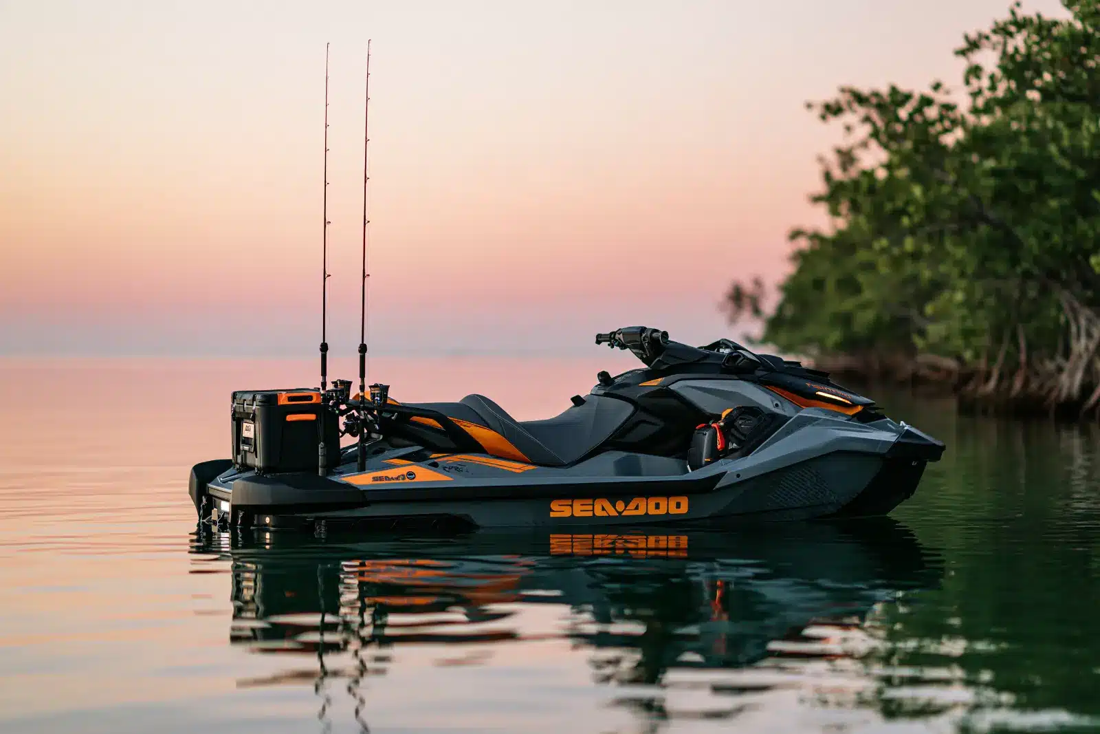 A gray and orange Sea-Doo FishPro with a cooler and two fishing rods mounted on the rear deck, floating on calm water beside mangroves at dusk