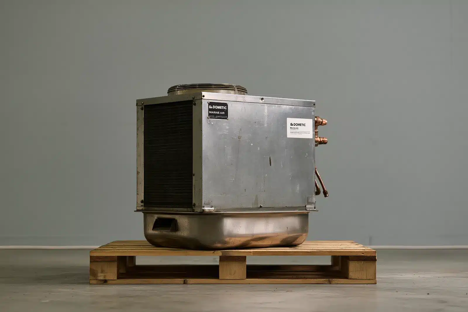 A used Dometic Marine Air self-contained air conditioning unit with its stainless drain pan and copper water fittings, sitting on a wooden pallet in a shop