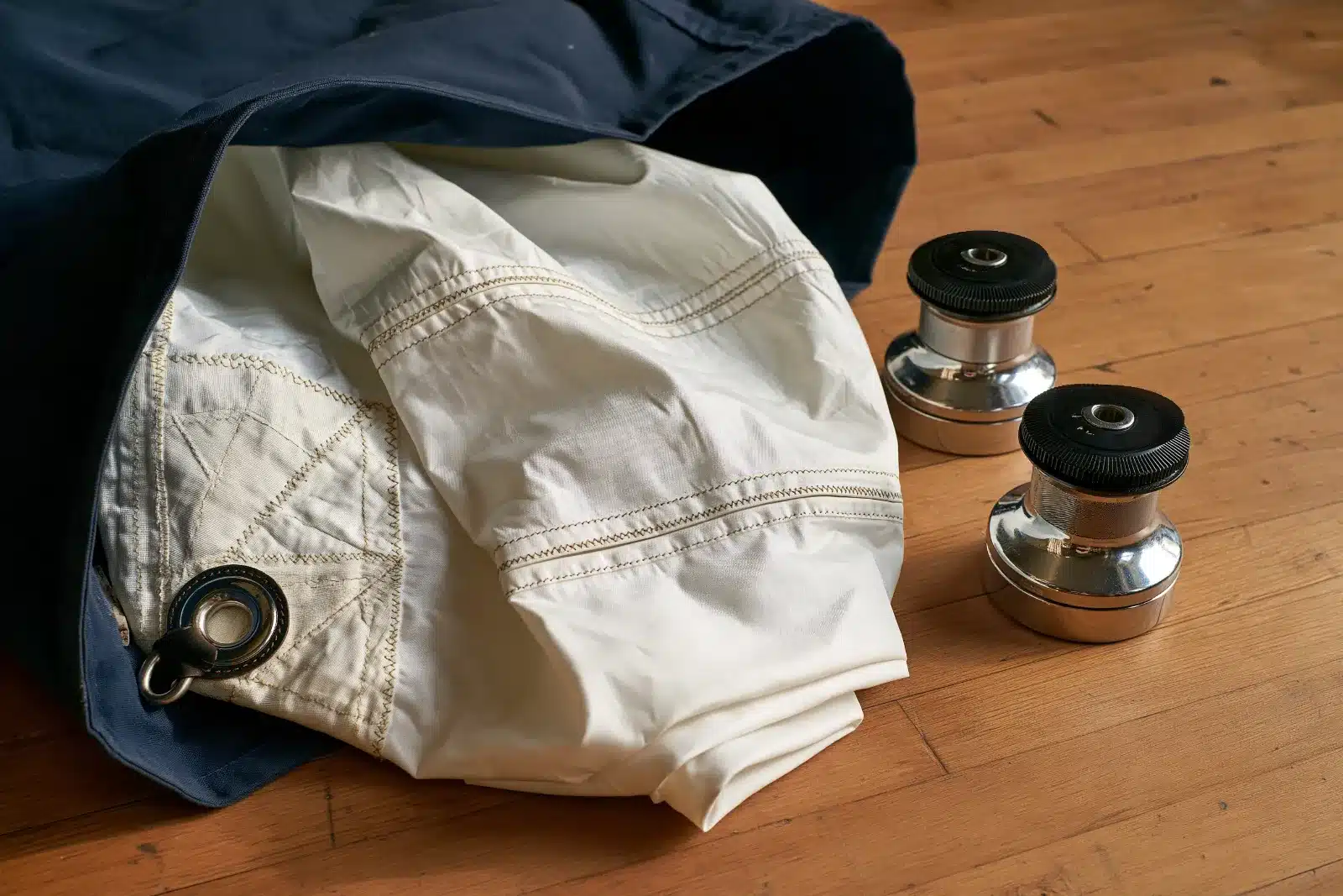 A used white Dacron mainsail with a stitched corner patch and stainless ring sliding out of a navy sail bag on a wooden floor, two chrome self-tailing winches beside it