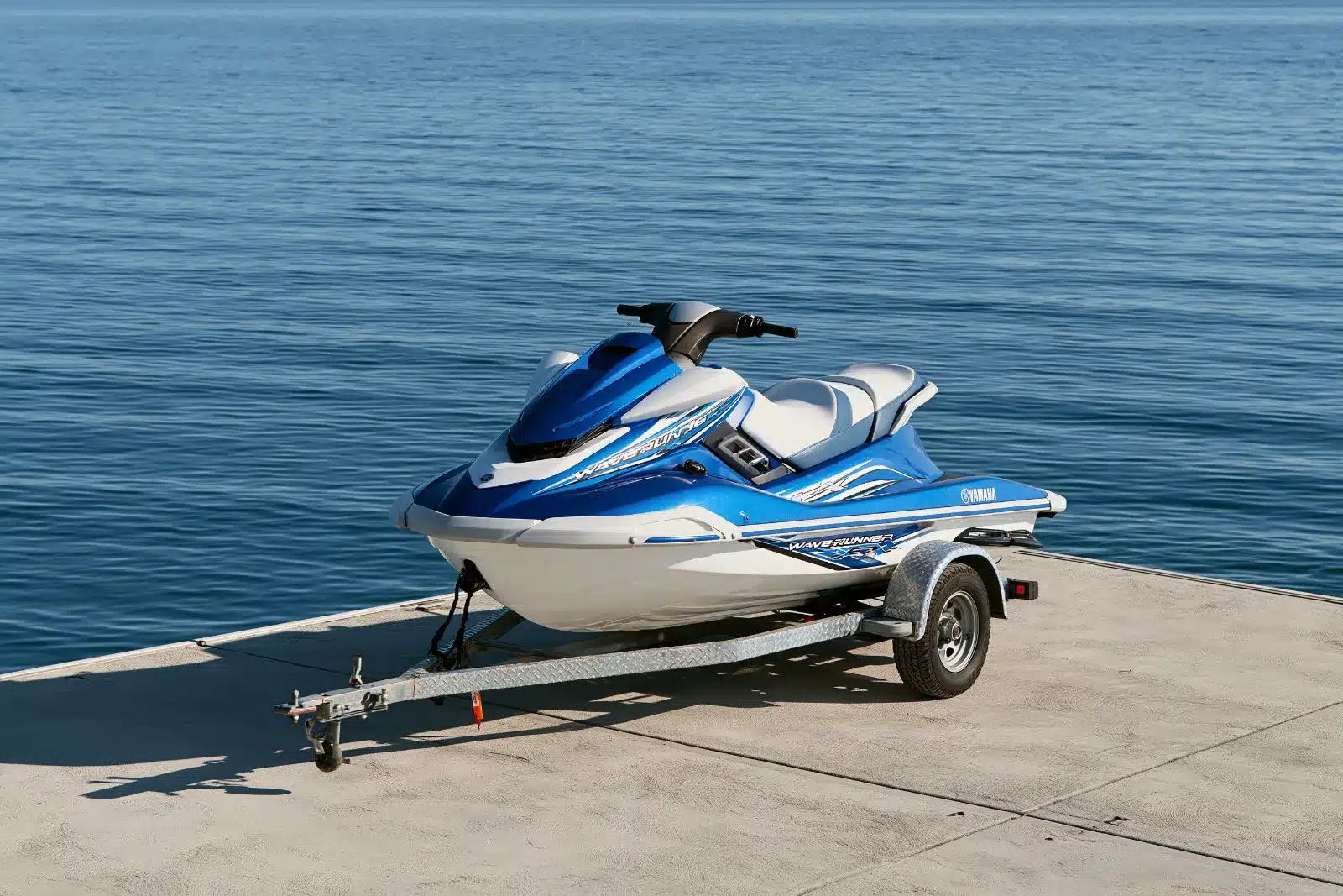 A blue and white Yamaha WaveRunner on a single-axle galvanized trailer, winch strap to the bow, parked on a concrete pad beside open blue water